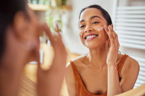 A woman happily applies EltaMD sunscreen on her cheek while looking in a bathroom mirror.